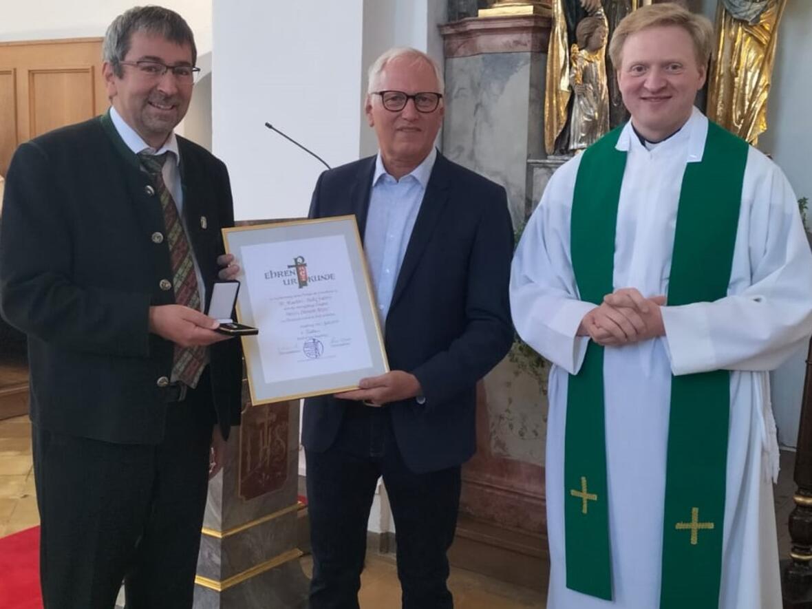 Drei Personen im Kirchenraum, Simon Peter hält eine Urkunde, die anderen gratulieren zum Jubiläum.