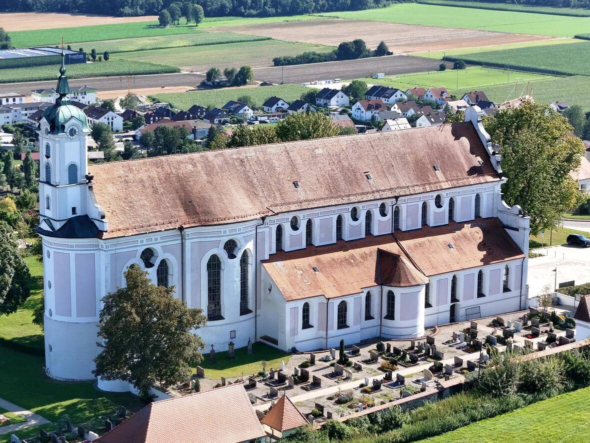 Kirche mit Grabsteinanlage im Vordergrund, umgeben von Feldern und Wohnhäusern.